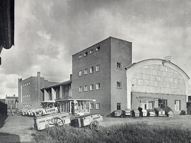 Gezicht op de voor- en achtergevel van het gebouw van de broodfabriek Lubro Bakkerij (Hogenoord 1) te Utrecht. Op de voorgrond enkele broodkarren.